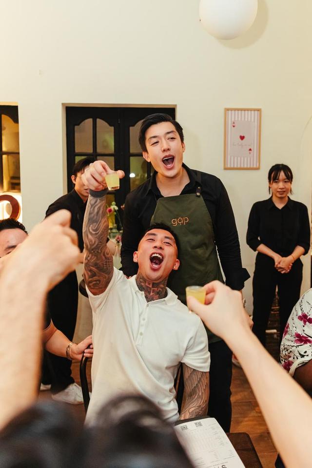 A gặp host in an olive apron cheering as guests raise shot glasses