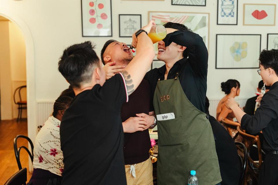 A gặp host in an olive apron pouring a drink for a laughing guest