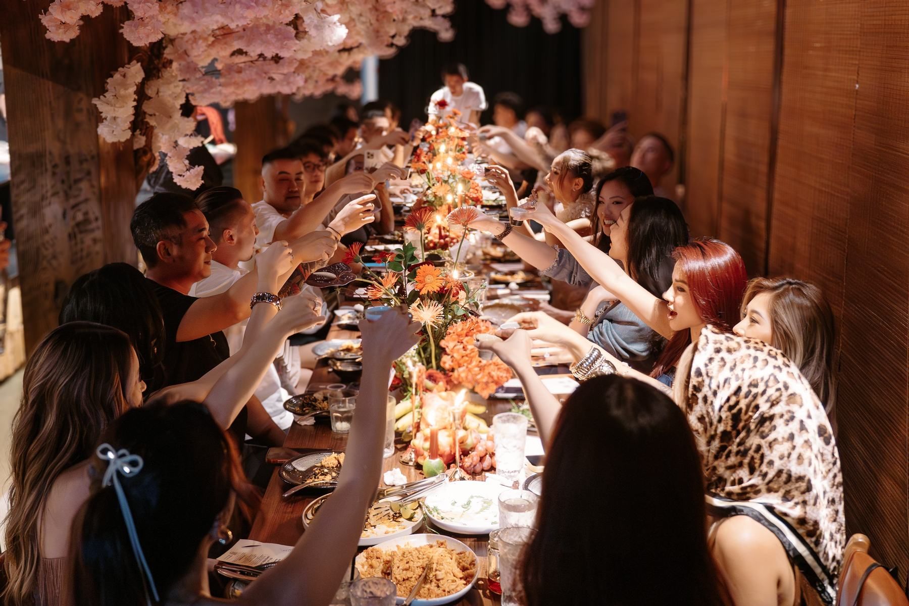 A long table of guests raising their glasses over orange flowers and candles